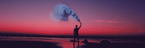 man stands on beach spraying blue gas in sunset sky