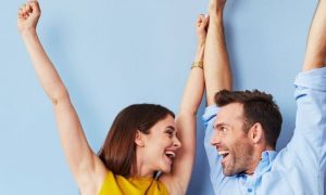 couple happily raises hands looking at each other blue wall