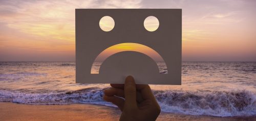 hand holding rectangle paper signaling sad face in front of beach waves in sunset