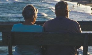 elderly couple sits on bench near beach facing backward enjoying cool weather watching people swimming