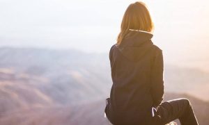 woman facing backward sits on mountain top looking at sunny sky