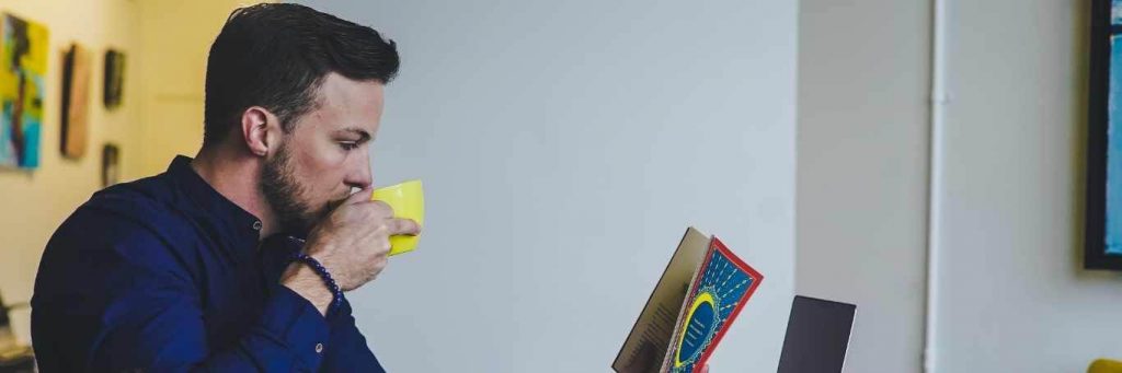 man sits in office drinking in yellow mug while reading book