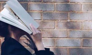 woman holds book covering face stands beside stone wall