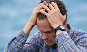 man wearing stripped shirt sits beside blue ocean scratching hair looking depressed anxious