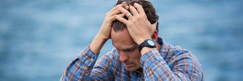 man wearing stripped shirt sits beside blue ocean scratching hair looking depressed anxious