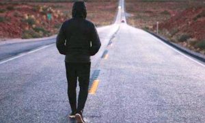 man wearing black hoodie facing backward walks alone on road between mountain