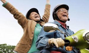 elderly couple rides scooter happily laughs while woman sitting behind raising hand
