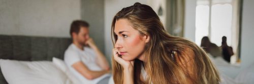 woman sits on bed thinking while man sitting hand in face in corner feeling unhappy