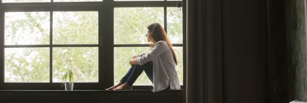 woman sits alone on bench beside window curtain looking outside