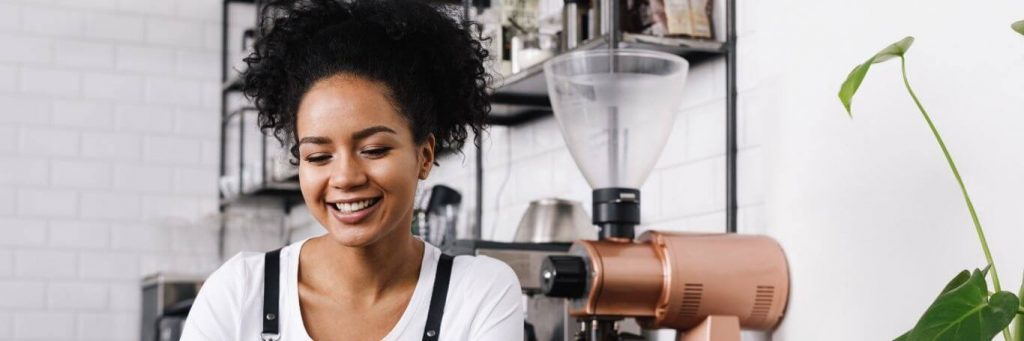 curly black hair woman sits in coffee shop beside coffee machine black shelf happily smile