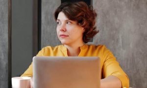 woman wearing yellow long sleeved shirt sits working on laptop looking aside