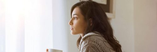woman sits alone in silent room drinking coffee looking through window