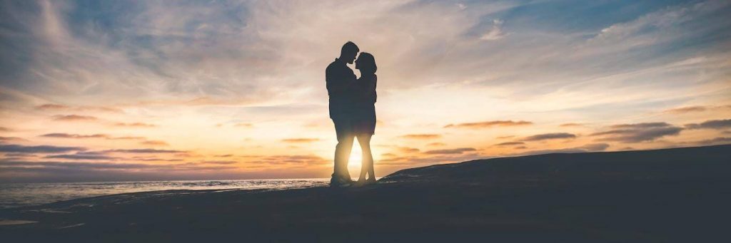 couple stands on rock beside beach hugging in beautiful sunset sky