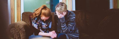 young woman sits on couch face down crying beside mom sitting beside hand in face crying