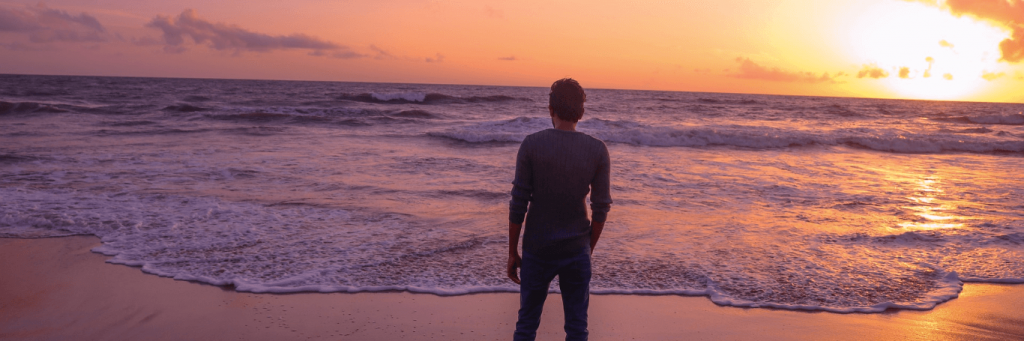 man facing backward stands hand in pocket on beach in beautiful sunset sky