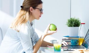woman sits on chair holding green apple while working on laptop