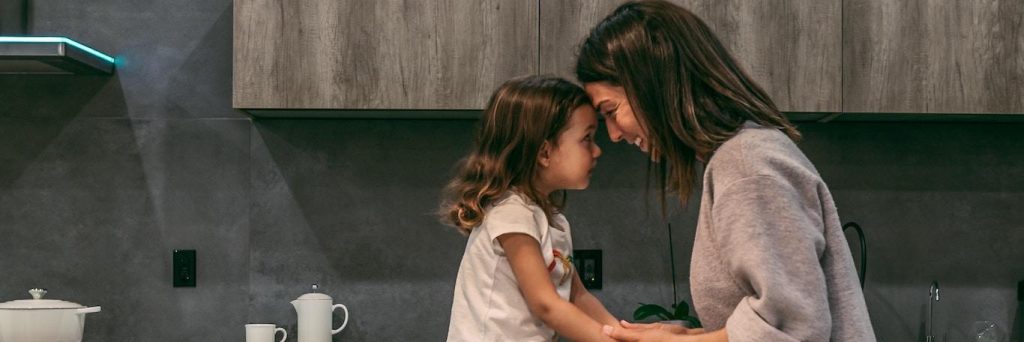 woman stands head to head with adorable daughter looking smiling in kitchen