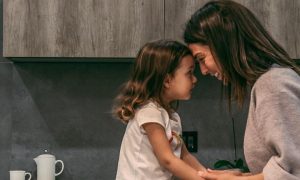 woman stands head to head with adorable daughter looking smiling in kitchen