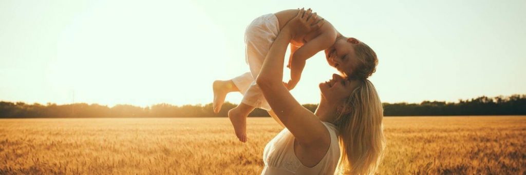 woman stands on field carrying little adorable son in sunny sky