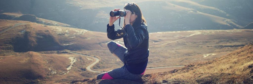 woman sits on rock seeing mountain world through binocular vision