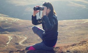 woman sits on rock seeing mountain world through binocular vision