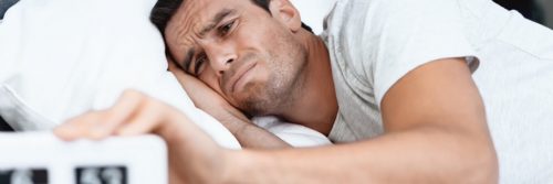 man lies on bed holding clock having insomnia problem