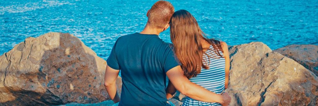 couple facing backward sits on rock side by side hugging looking at blue ocean
