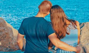 couple facing backward sits on rock side by side hugging looking at blue ocean