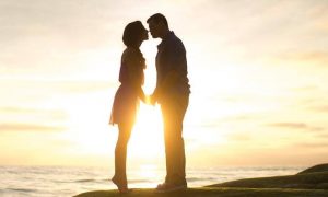 couple stands on rock beside ocean kissing in beautiful sunset