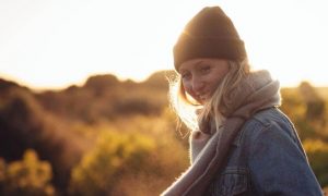 woman wearing black hat purple scarf stands in field happily smiling in sunny sky