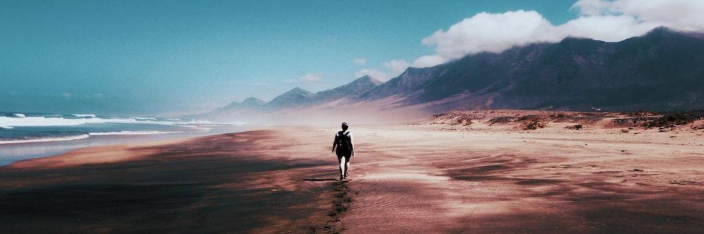 person walks on sand beside blue ocean mountain blue cloudy sky