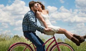 man rides biking carrying girlfriend while woman sitting in front kissing in blue cloudy sky