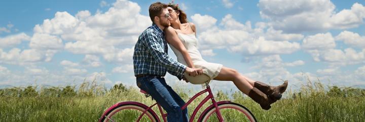 man rides biking carrying girlfriend while woman sitting in front kissing in blue cloudy sky