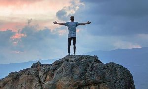 man stands on rock facing backward gratitude life in blue cloudy sky