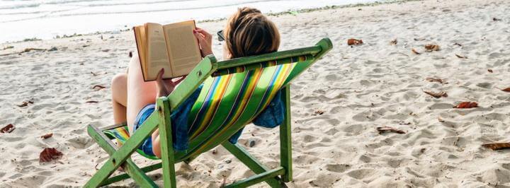 woman wearing sunglasses sits on chair reading book on beach