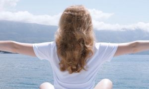 woman sits beside blue ocean facing backward gratitude life in cloudy sky