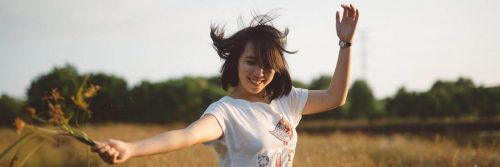 short hair woman dances on field happily smiling carrying flowers in clear sky