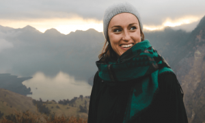 woman wearing winter hat scarf stands smiling on top of mountain in foggy weather