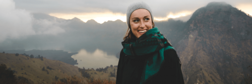 woman wearing winter hat scarf stands smiling on top of mountain in foggy weather