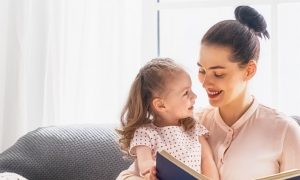 woman sits on couch smiling reading book for little adorable daughter in living room