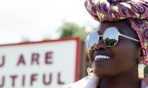african woman wearing sunglasses stands beside street board happily smiling