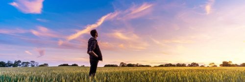 man stands alone in field hands in pocket looks at beautiful partially cloudy blue sky