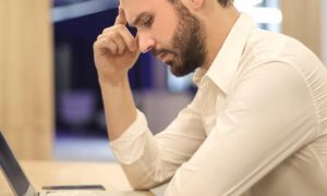man sits in library hand in forehead thinking looking at laptop