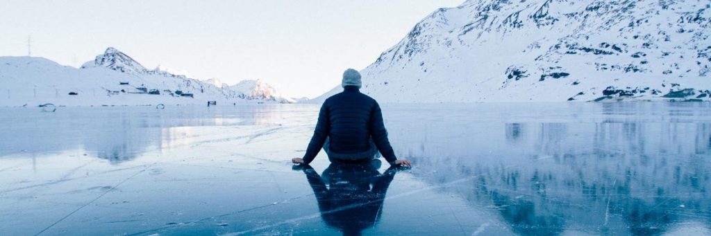 man facing backward sits alone on iceberg looking at snowy mountain clear sky