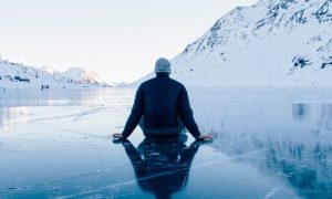 man facing backward sits alone on iceberg looking at snowy mountain clear sky