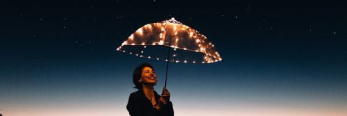 elderly woman stands holding umbrella filled up with lights happily smiling in dark star sky