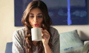 woman sits on couch holding white mug blowing