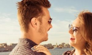 couple wearing sunglasses stands near blue ocean laughing